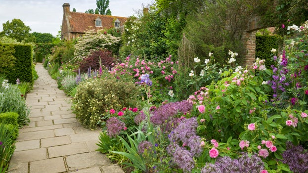 Pink and white roses, lilac and purple alliums, and other purple, pink and white flowers bloom in a flowerbed next to a stone path, with the house in the background, at Sissinghurst Castle Garden, Kent