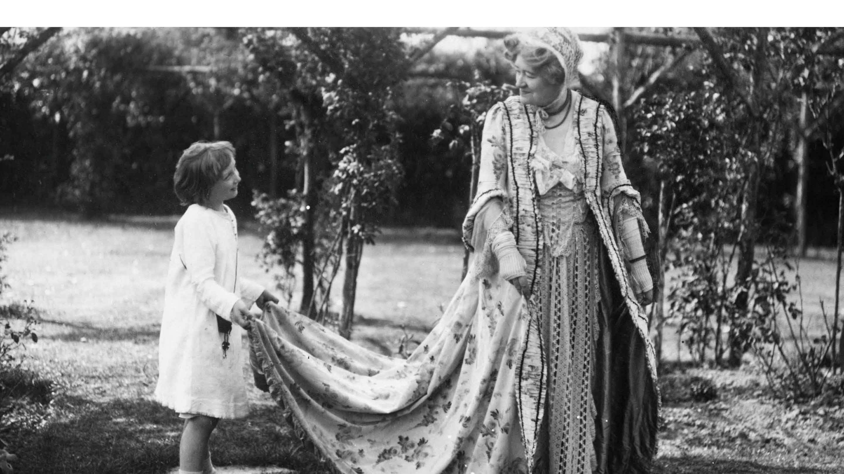 Black and white archive photograph of Ellen Terry in costume with her grandchild Nellie Craig holding the train of her costume, at Smallhythe Place.