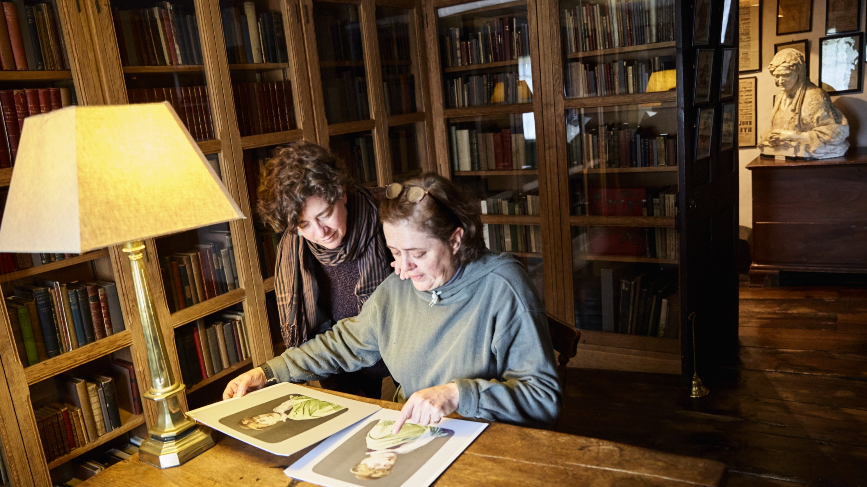 Two visitors leaning over a desk looking at some pictures. In the background are wood and glass cases with books in.