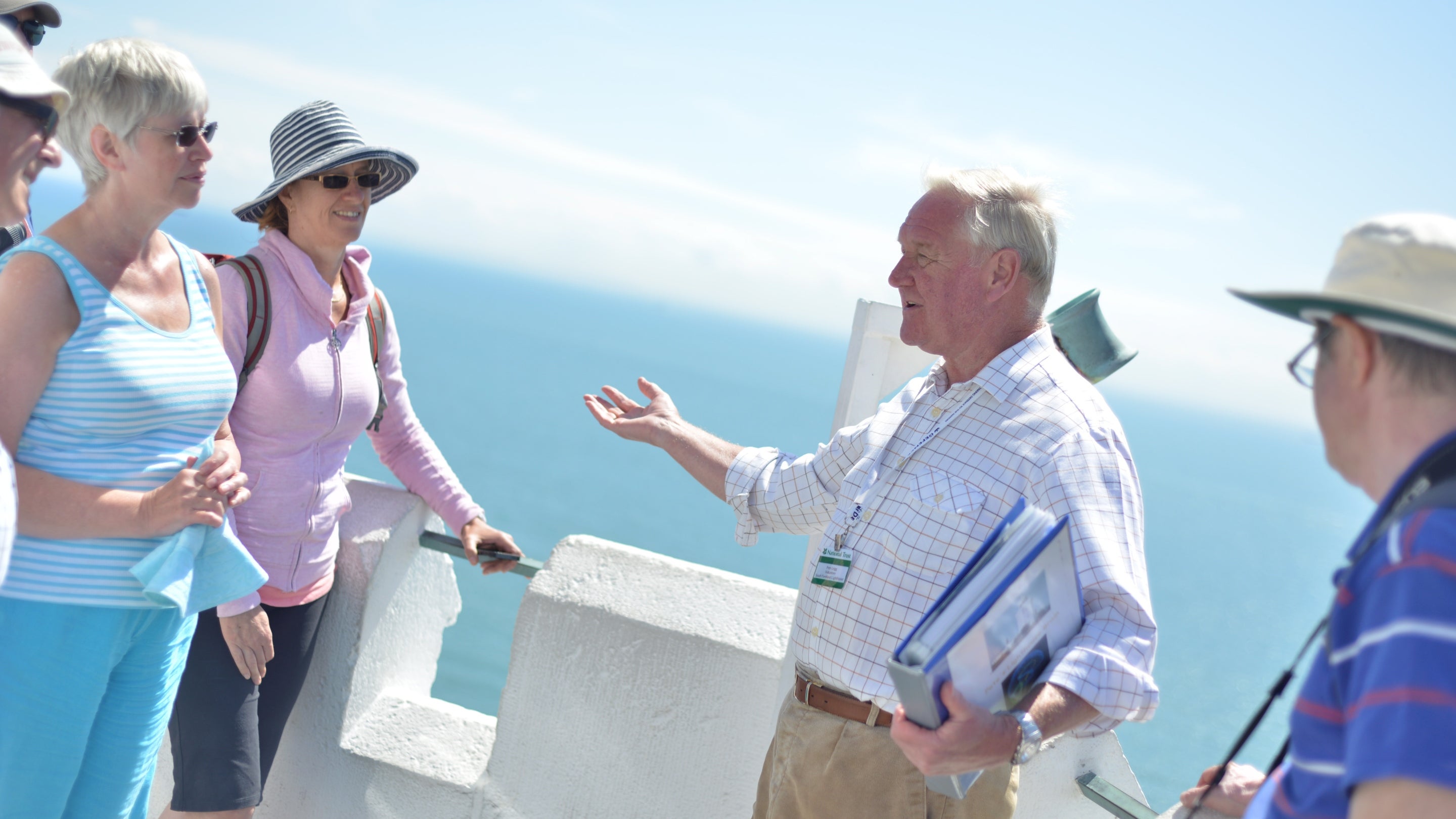 A group of people in summer clothing listen to a volunteer guide speak, on the balcony at the top of South Foreland Lighthouse in Kent.