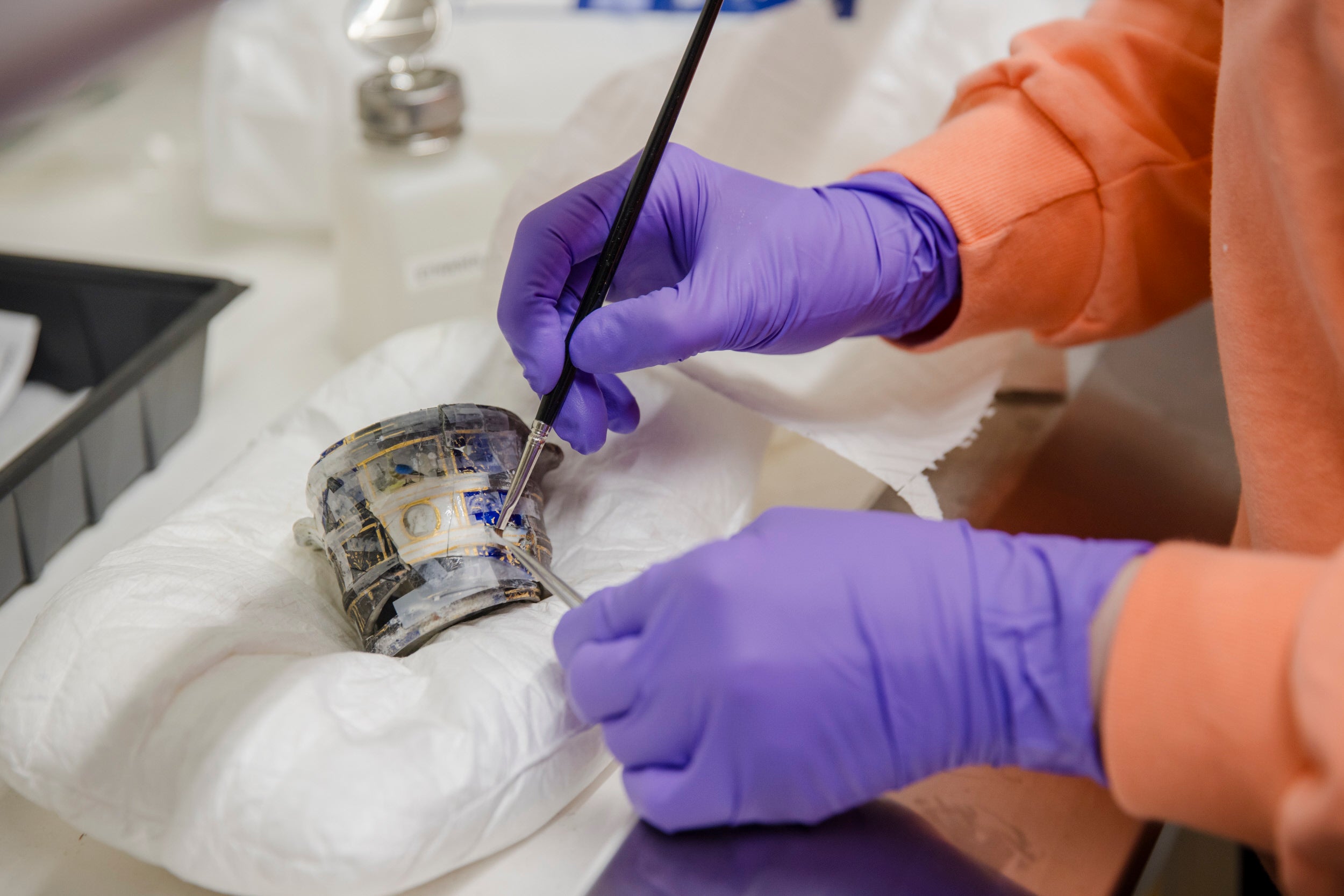 Someone wearing purple gloves holding a tools in both hands as they work on a piece of blue and white ceramic
