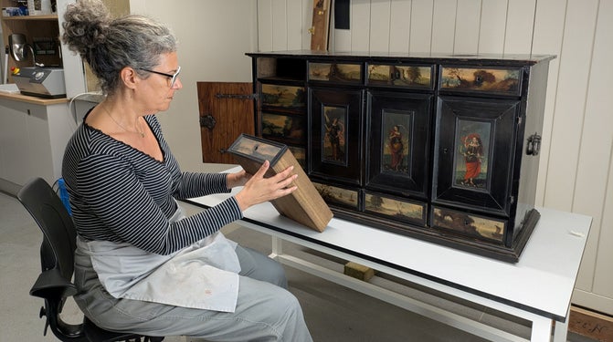 A person sitting at a table holding and looking at a wooden drawer, with a decorated black wooden cabinet on the table in the background.