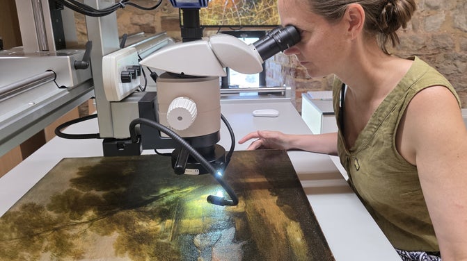 A person sitting at a table looking through a microscope which is positioned over a paintings