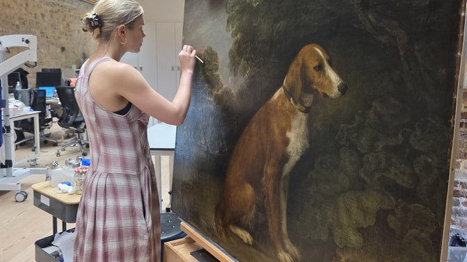 Person with a paintbrush in their hand, which they are holding up to a large painting of a dog that is positioned on an easel in front of them.