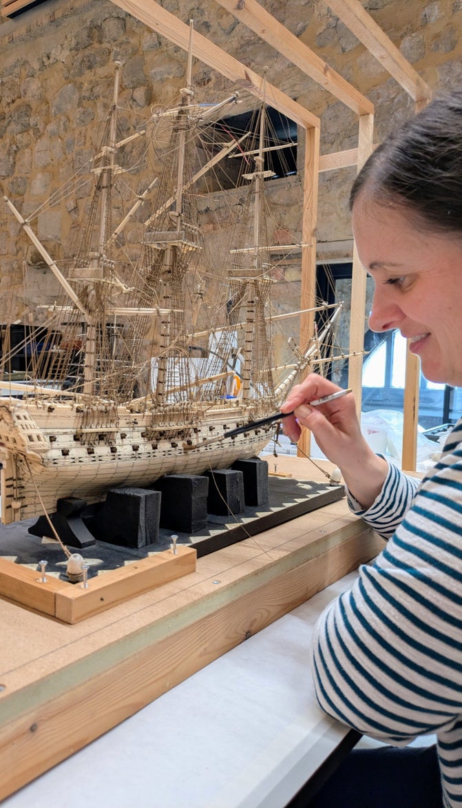 A person is sat at a desk wth a paintbrush in their hand that they are using on a wooden model of a ship that is positioned on the desk in front of them.