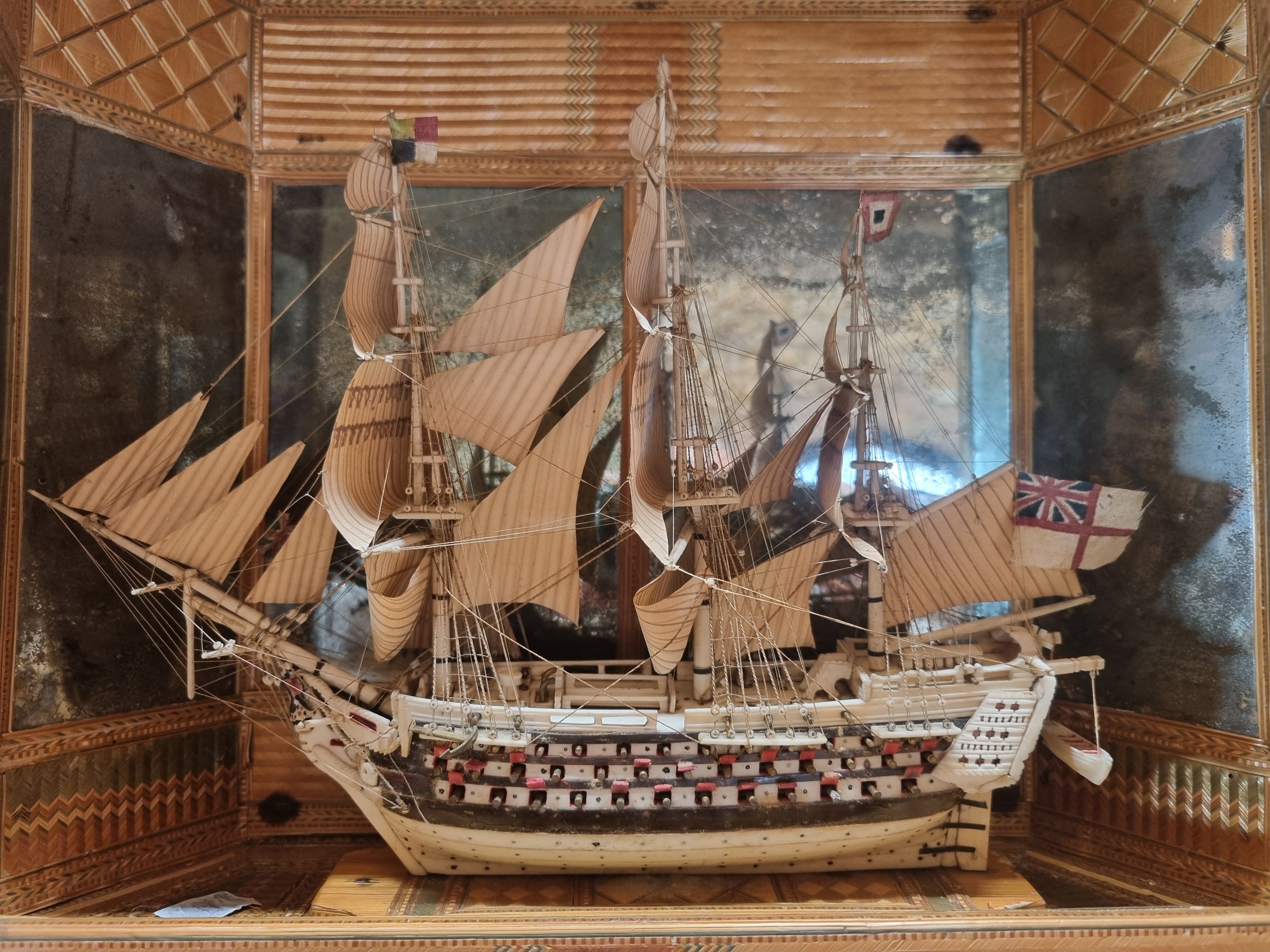 A model of a white ship with beige sails surrounded by a light brown case which is open at the front.