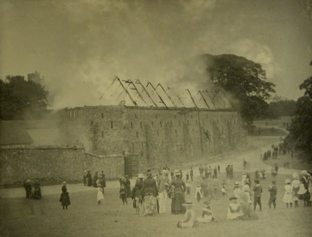 A black and white photo of a large barn building on fire. There is smoke coming out of the roof, of which there are only beams left. A crown of people are standing watching.