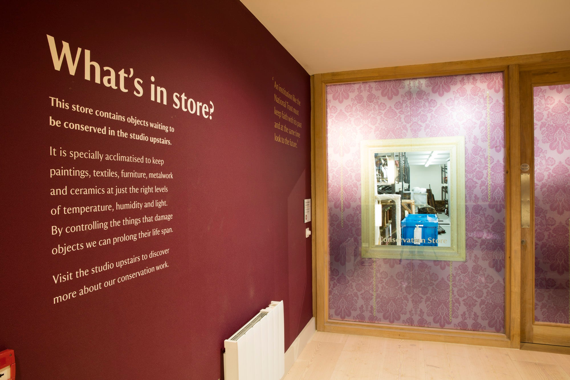 A dark red wall on the left with "What's in store?" in large white writing at the top above smaller writing. To the right is a large glass window and door. There is a pattern over most of the window, with a square in the middle that is clear, giving a view into the room on the other wide.