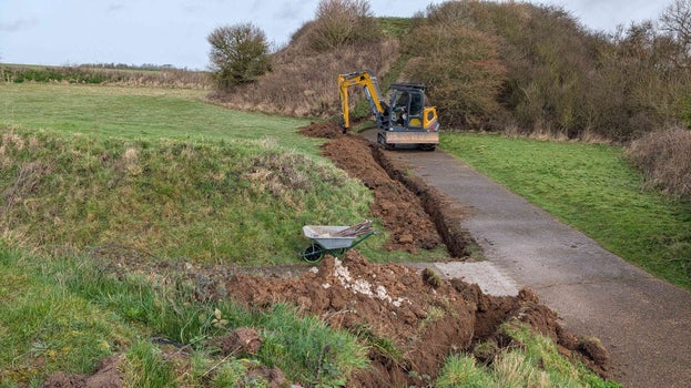 Image of a trench being excavated alongside a narrow road to a large building