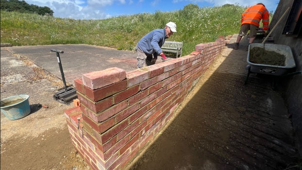 Image of people constructing a brick wall against a sloping path with embankments behind