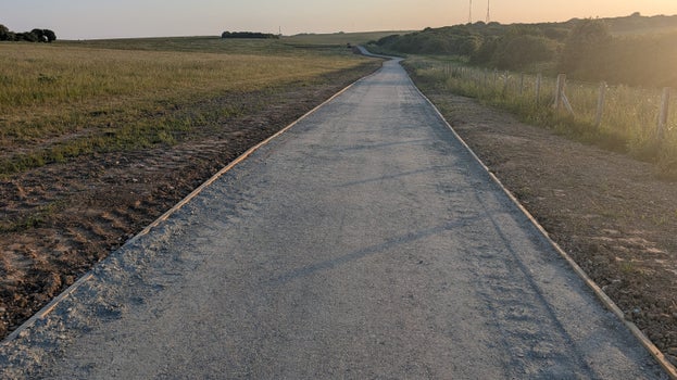 Image of a wide grey compacted path on a gentle slope with a setting sun's rays to the right of the camera