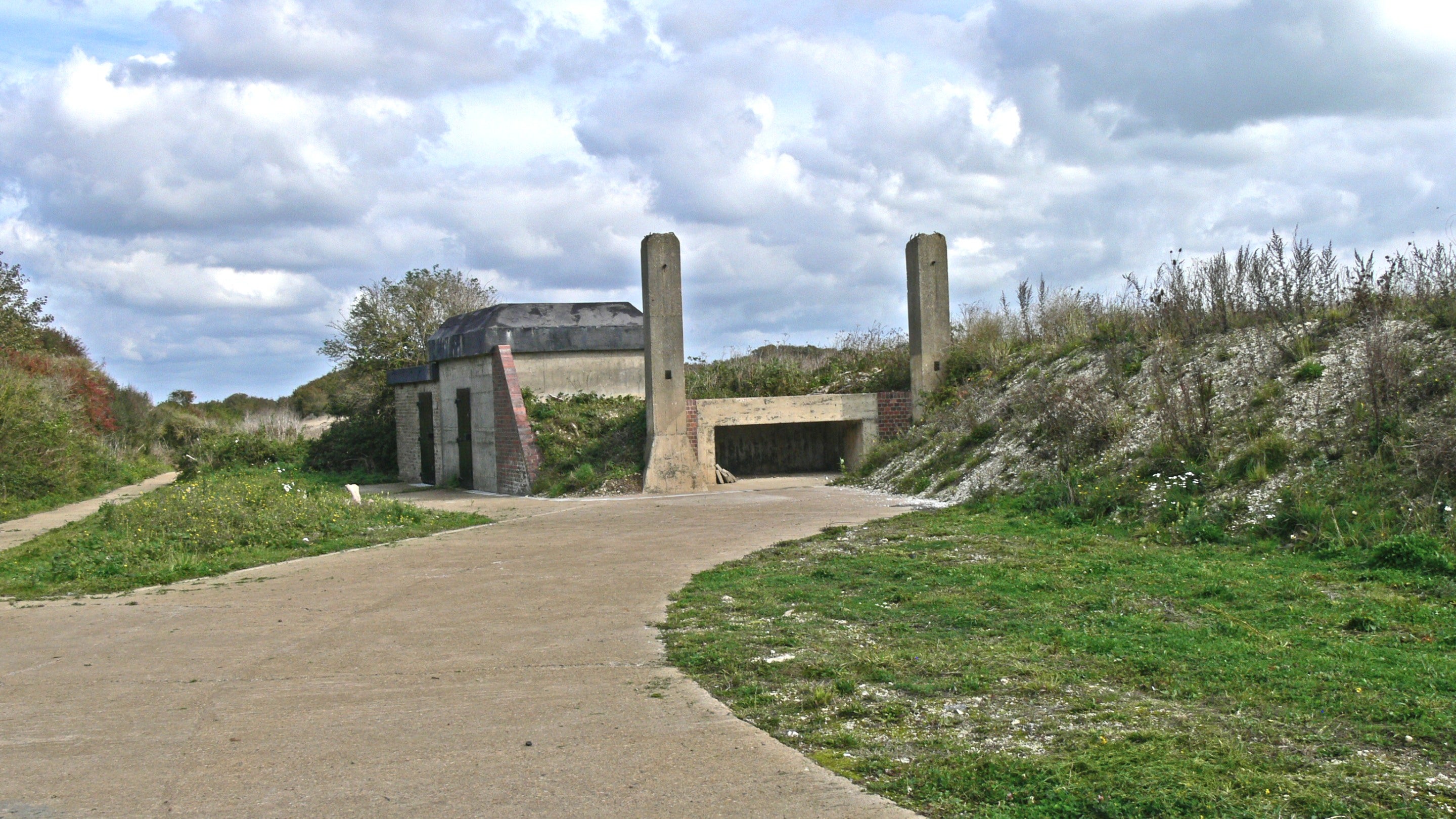 Image of a semi-circular concrete structure at ground level, with two buildings in the background on a sunny day with patchy cloud