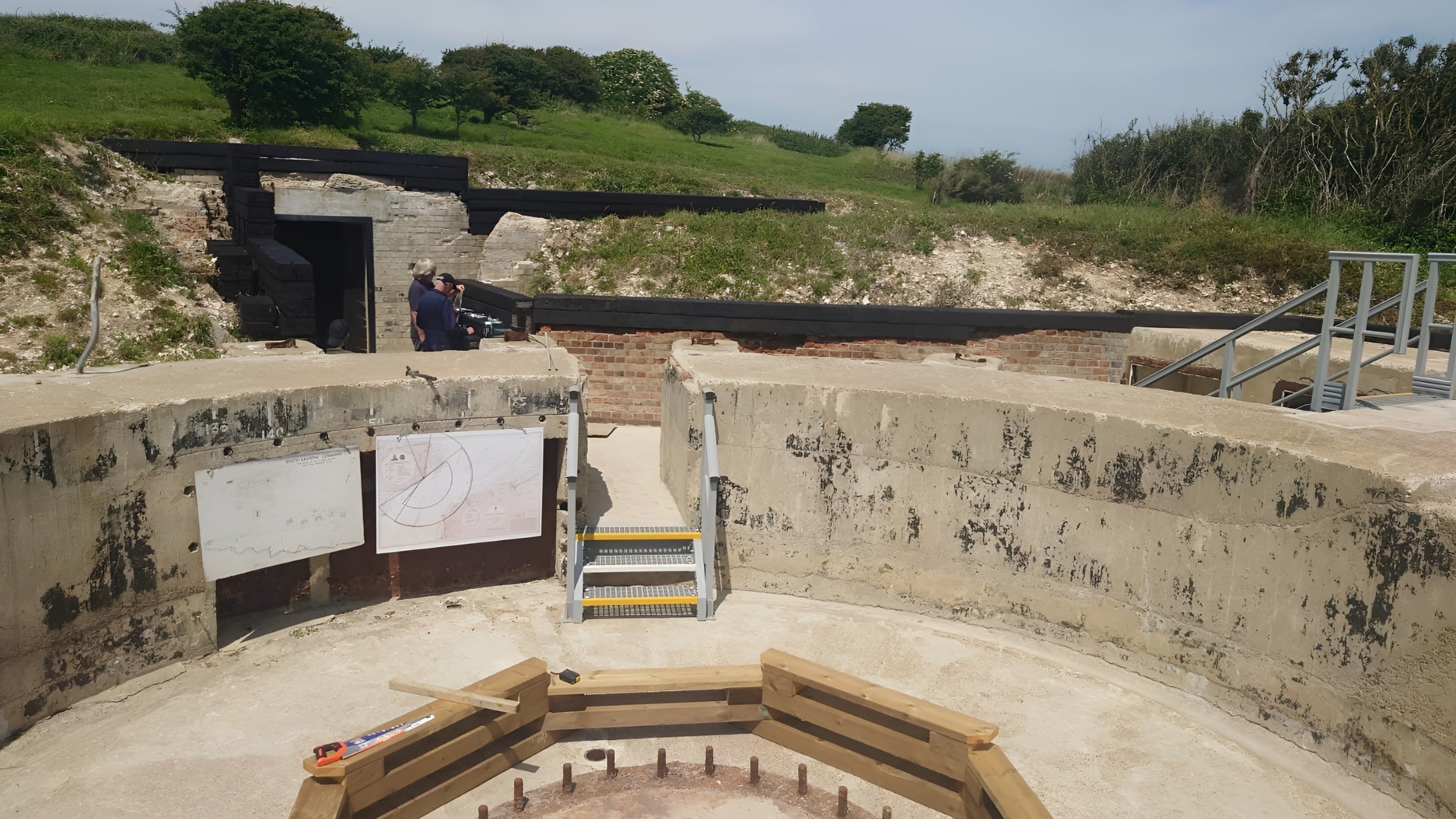Image of a circular concrete gun emplacement with the holding down bolts visible and with the visitor seating and interpretation boards in place