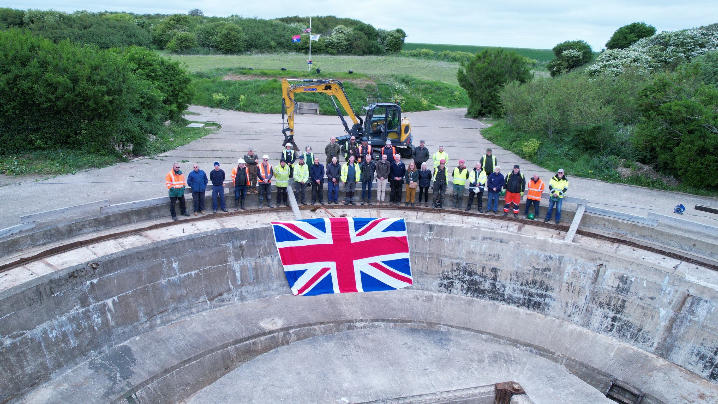 Image of a large group of people standing on the edge of a concrete pit with a Union Flag hanging into the pit with trees and grass in the background