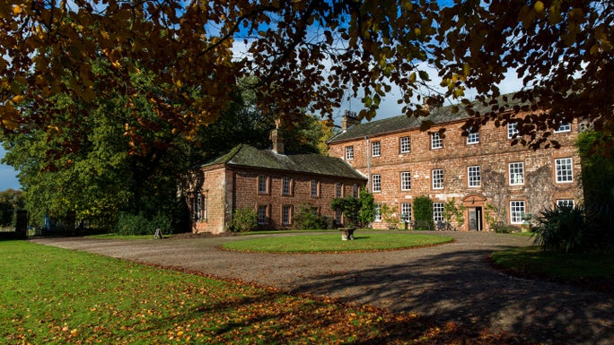 An old red-brick house with autumnal light shining on it and trees and vegetation to the left