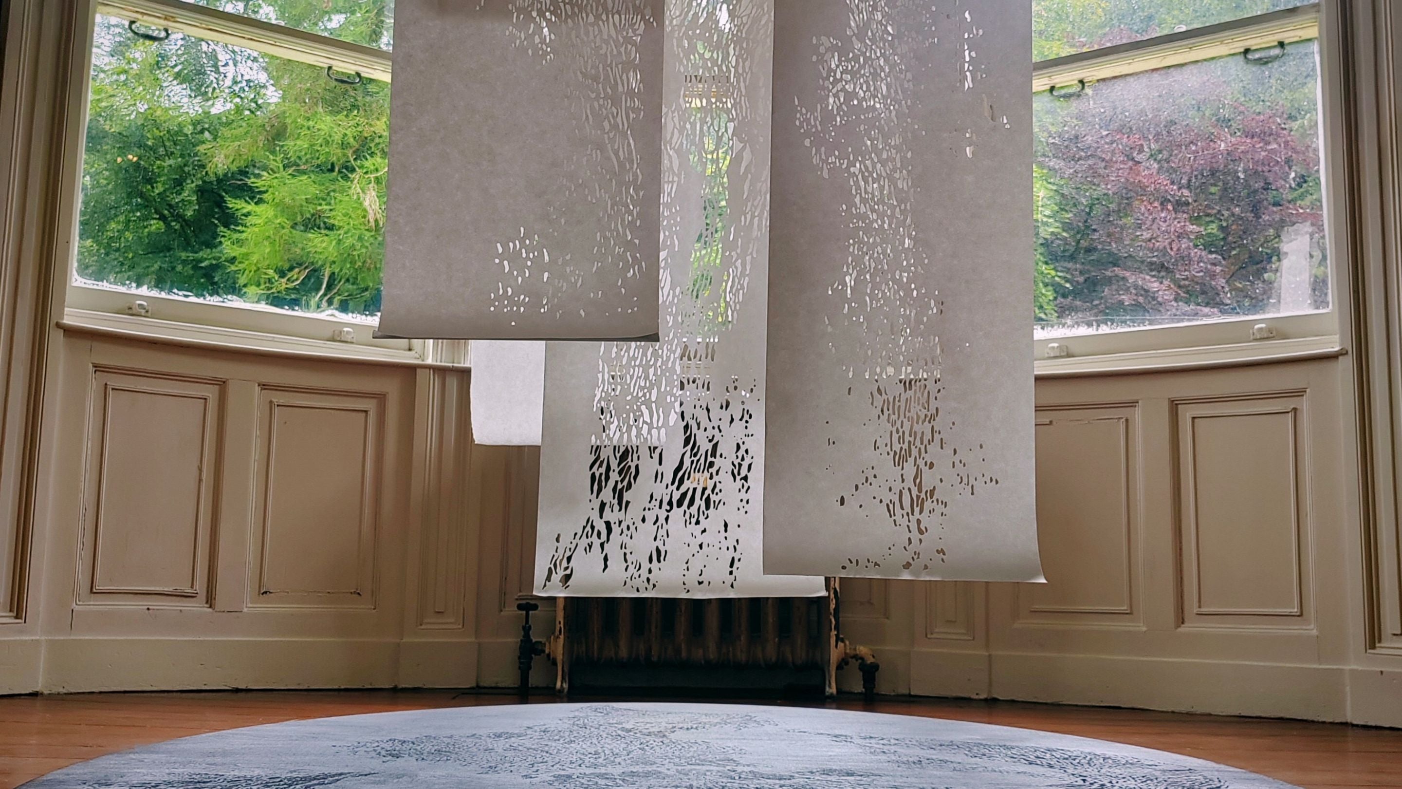 Three rolls of intricately cut white paper hang from the ceiling and float above a  dark circular platform in a room.