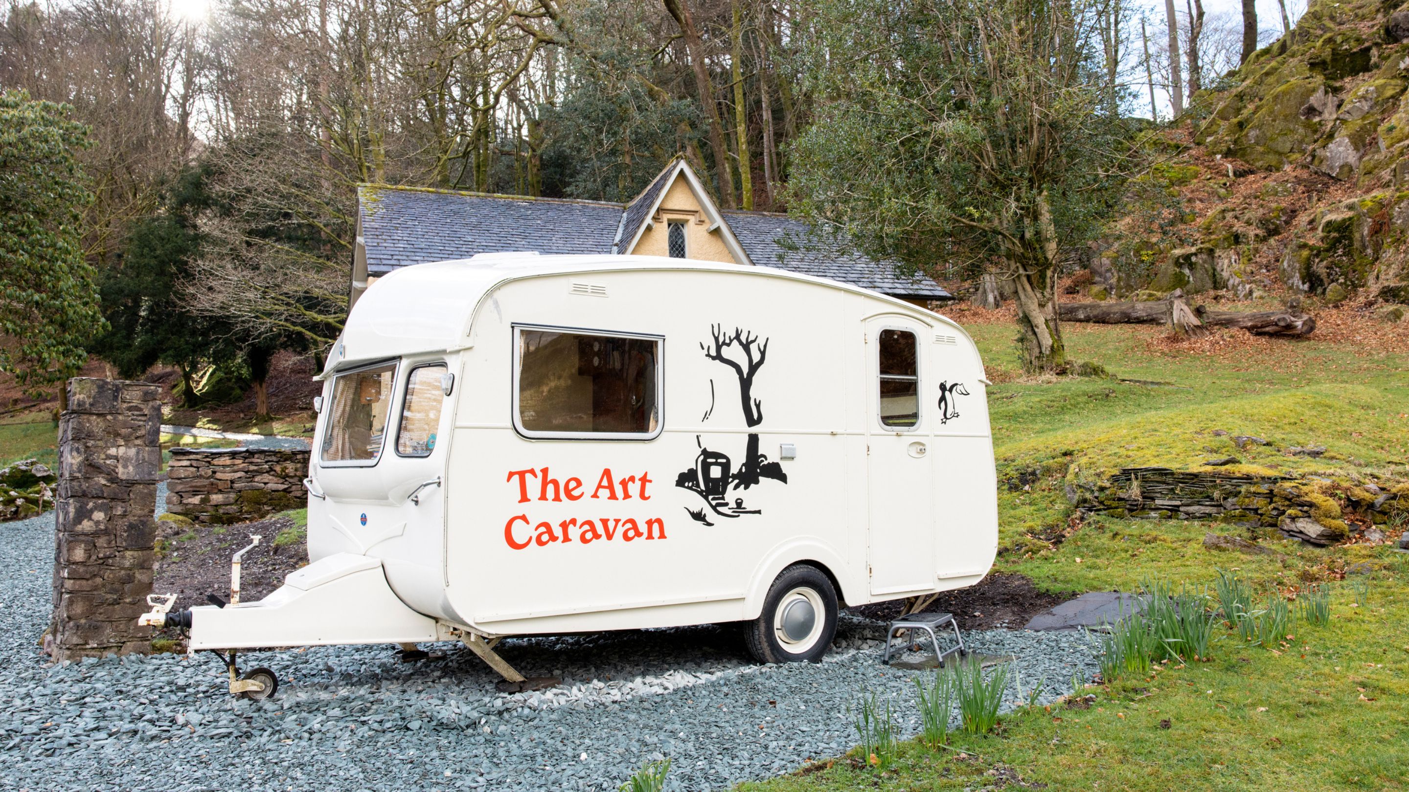White caravan labeled 'The Art Caravan' with illustrated designs, parked on a gravel path near a stone house in a wooded area at Allan Bank, Grasmere