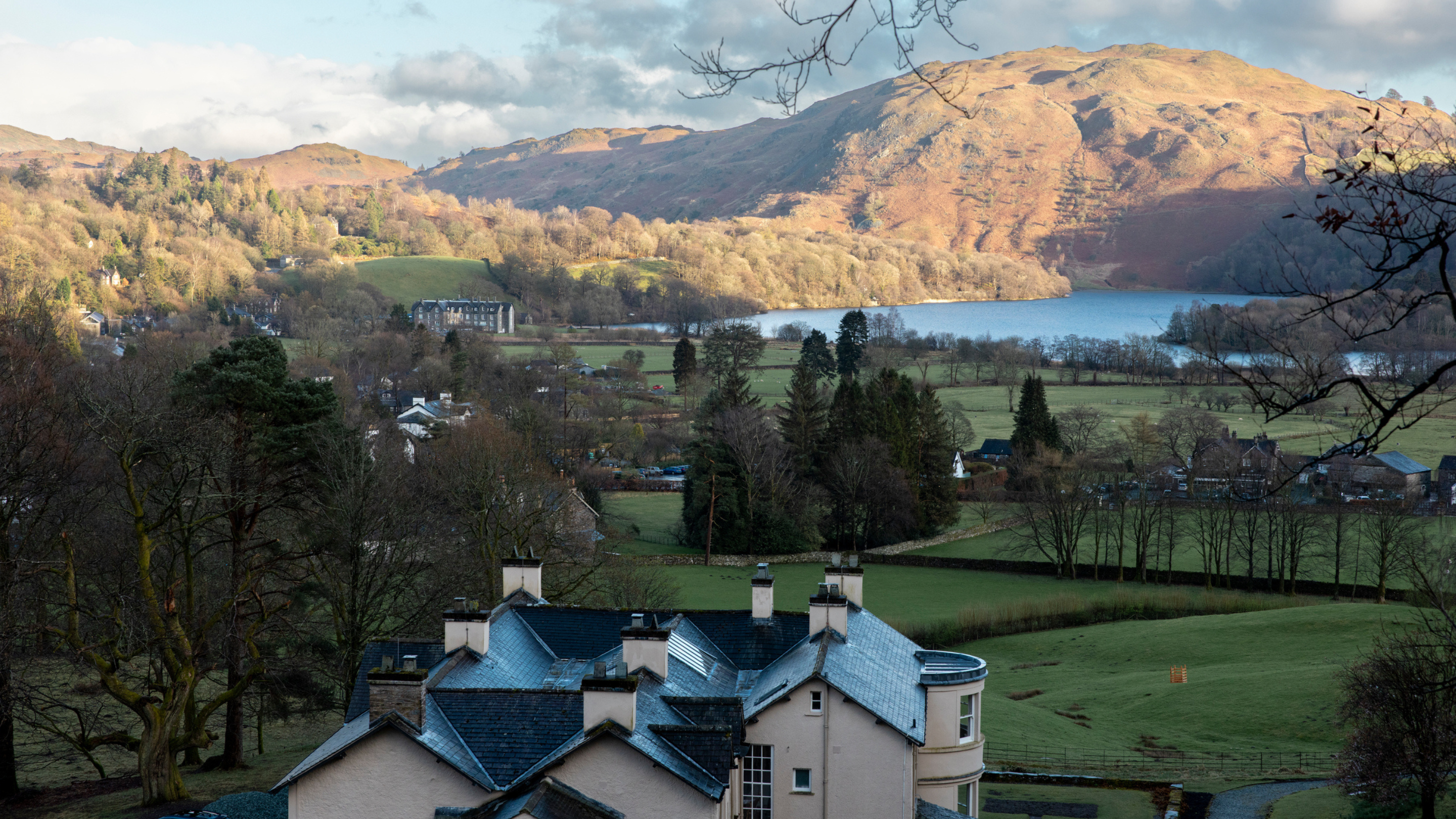 A historic house with slate roofs and chimneys surrounded by lush greenery, with a lake and sunlit hills in the background. A tranquil springtime woodland landscape.