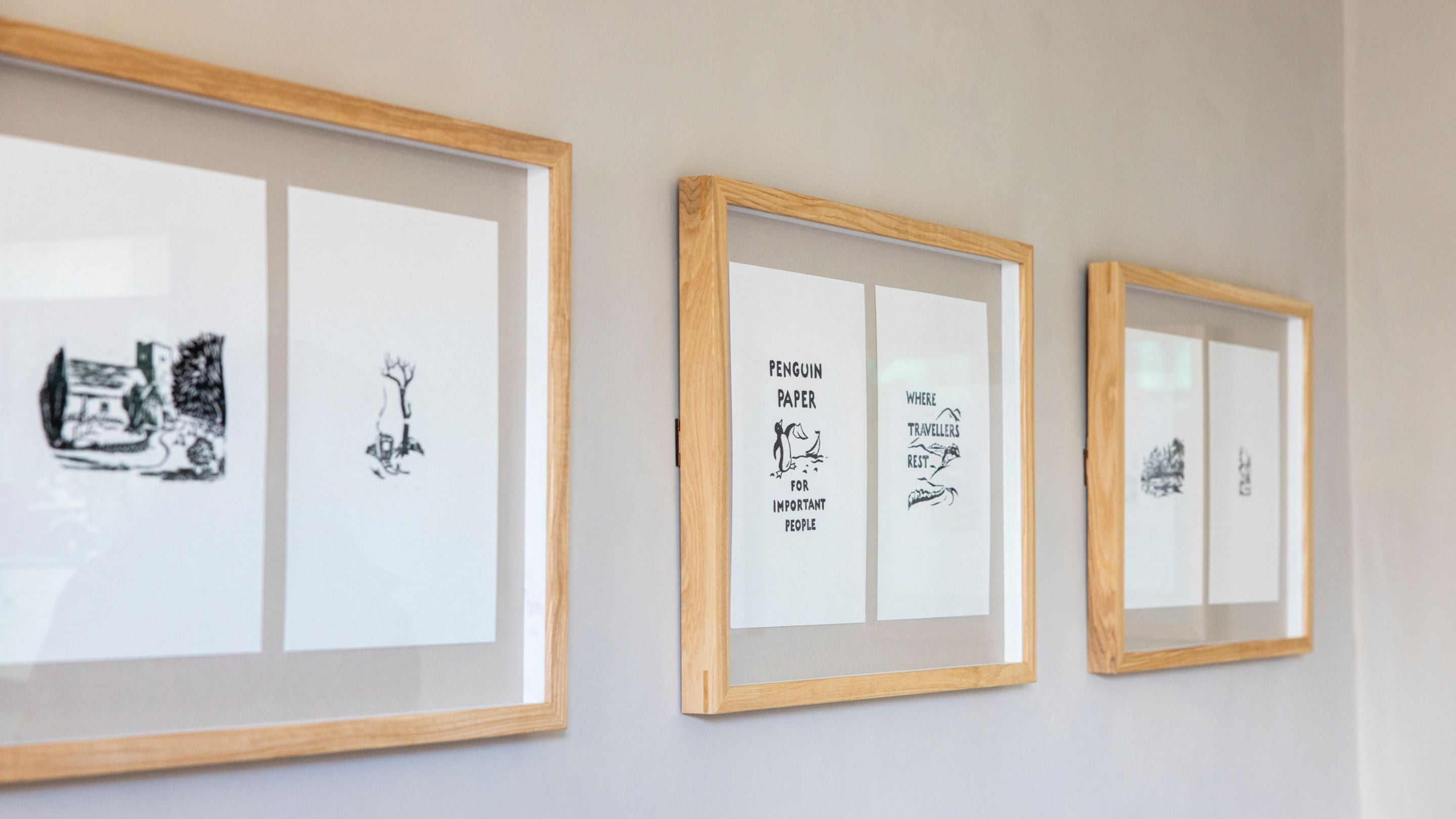 Three black and white prints framed in light wood are displayed in a line on a cream wall.