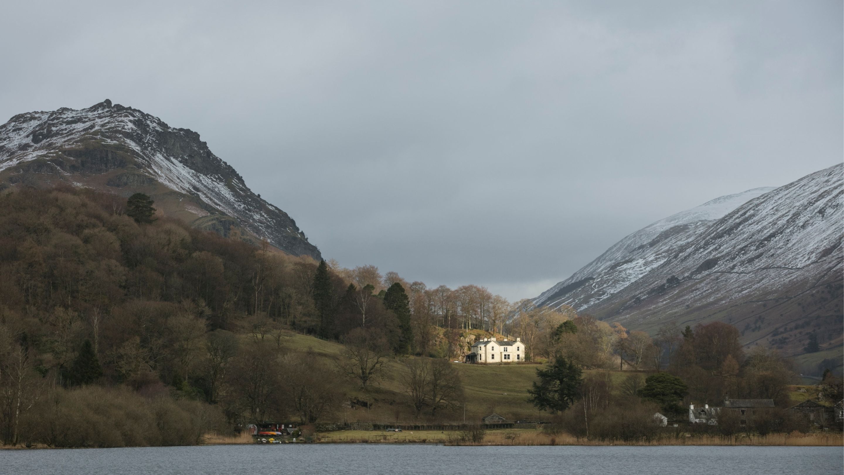 Winter scene of a house perched on hill, surrounded by higher fells and a lake in the foreground