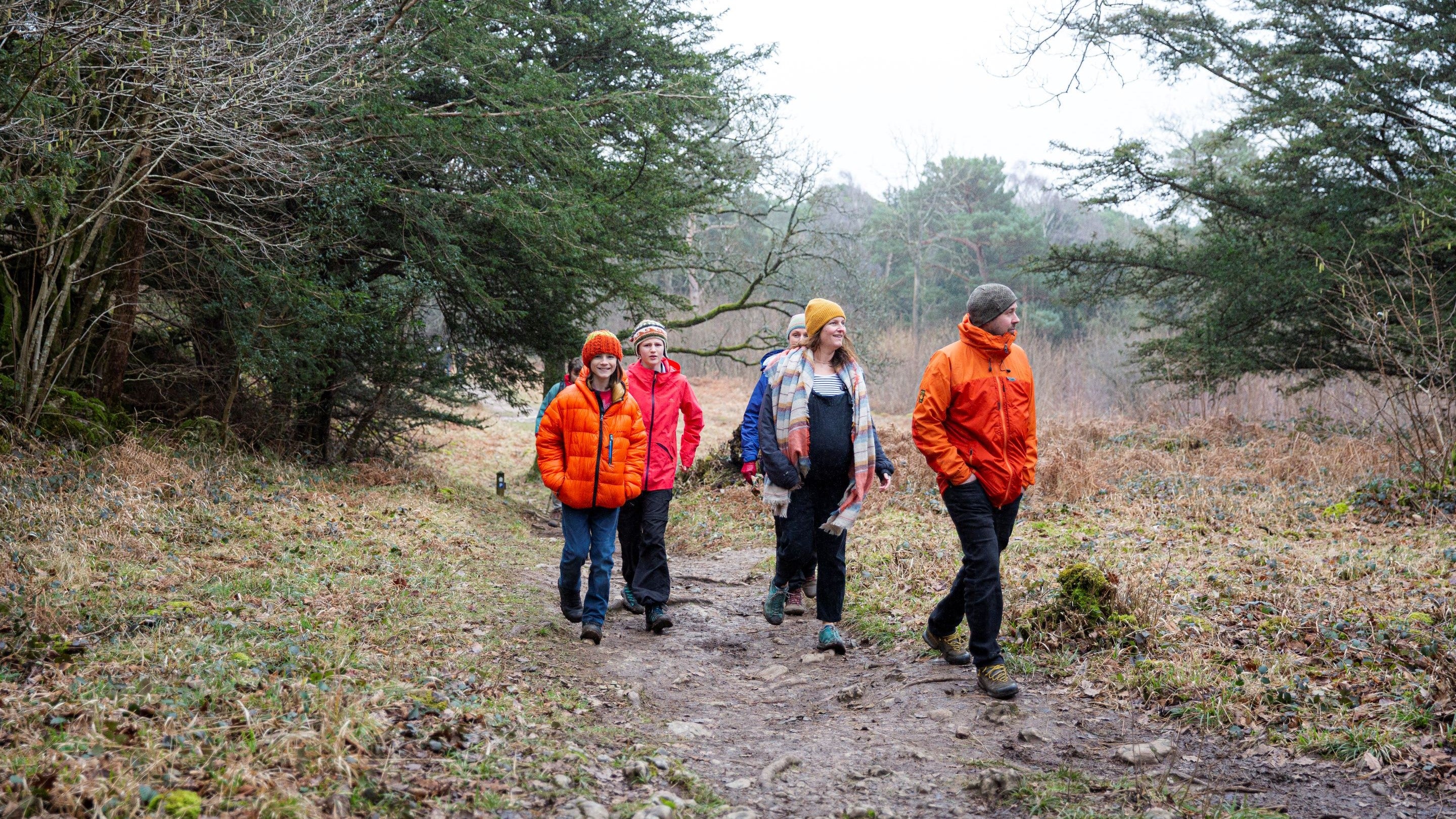 Family, dressed in coloured coats and hats for the cold, walk to the Pepperpot through the ancient wood at Arnside and Silverdale, Cumbria. It's a grey day and the ground is muddy.