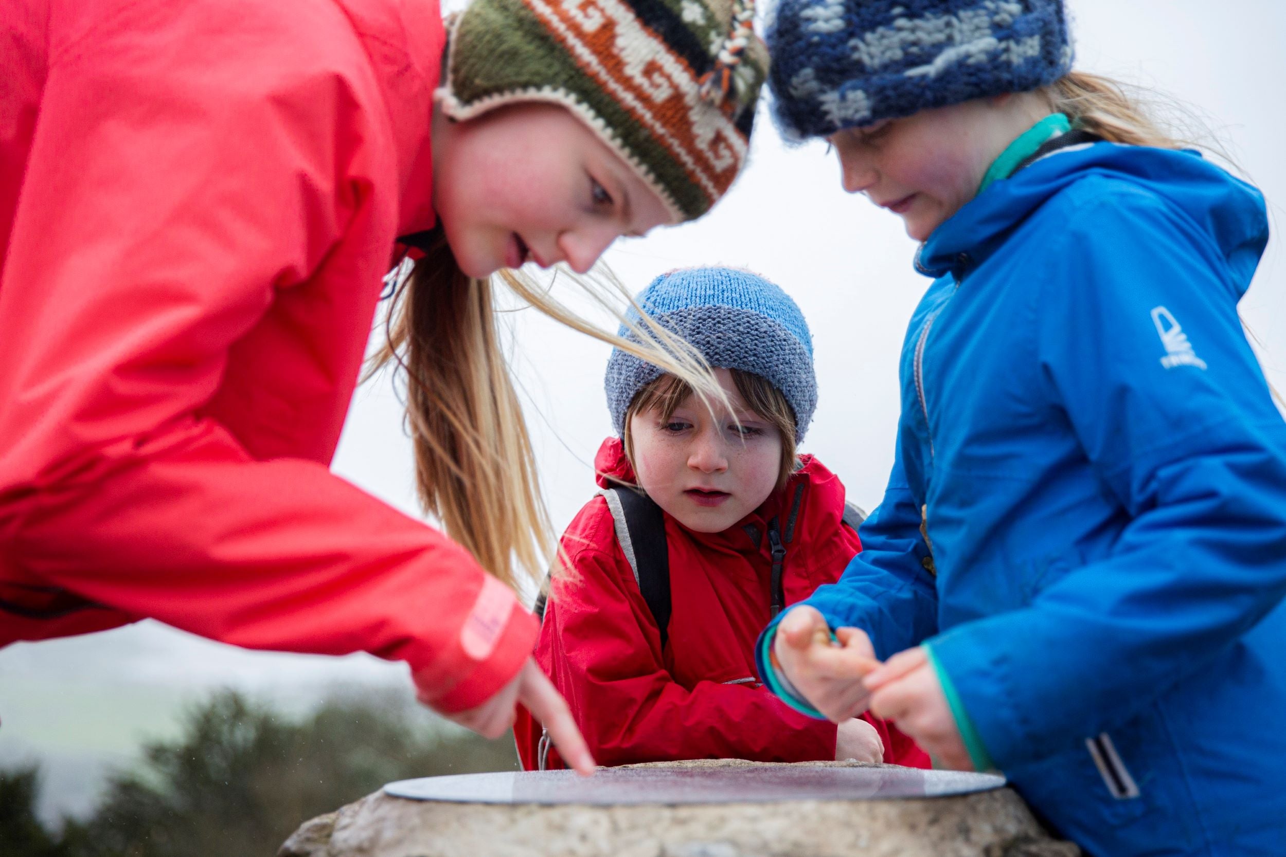 Three children looking at the Pepperpot monument at Arnside and Silverdale. They are all wearing brightly coloured waterproof coats and wooly hats.