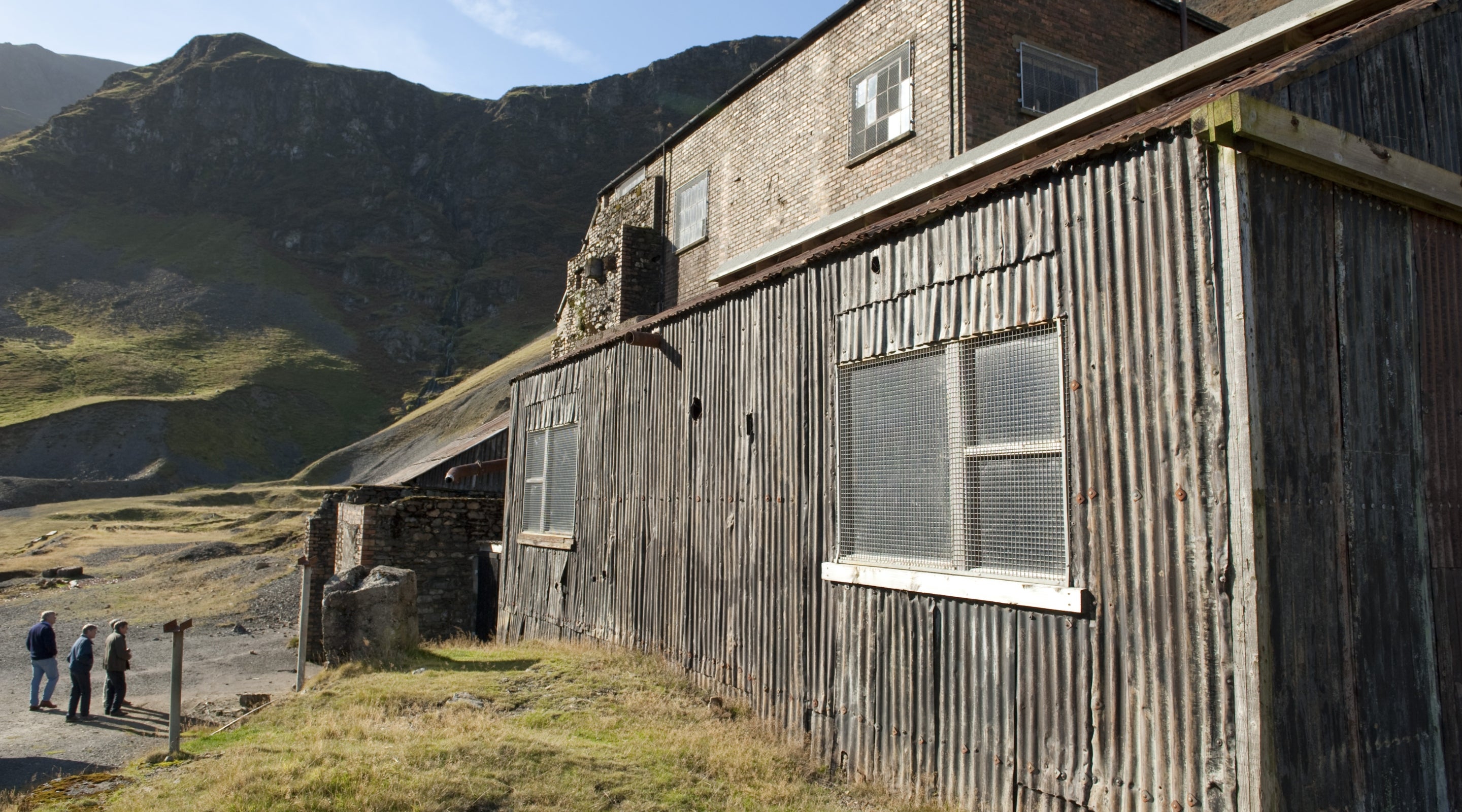 Visitors about to enter Force Crag Mine on a sunny day with the old building in the foreground and Coledale Valley behind