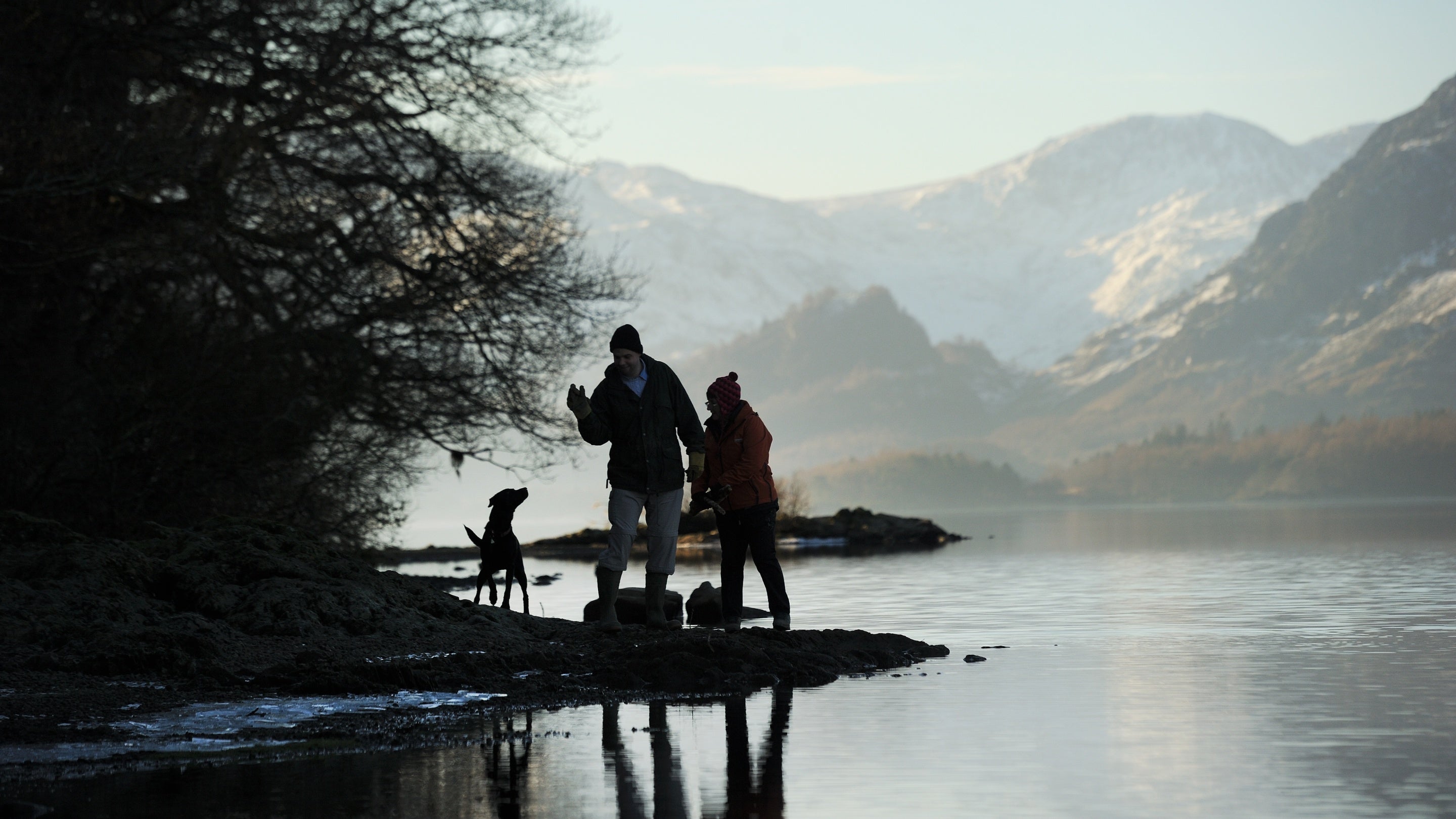 Two people and their dog by the shore of Derwent Water in winter, set against a mountainous backdrop