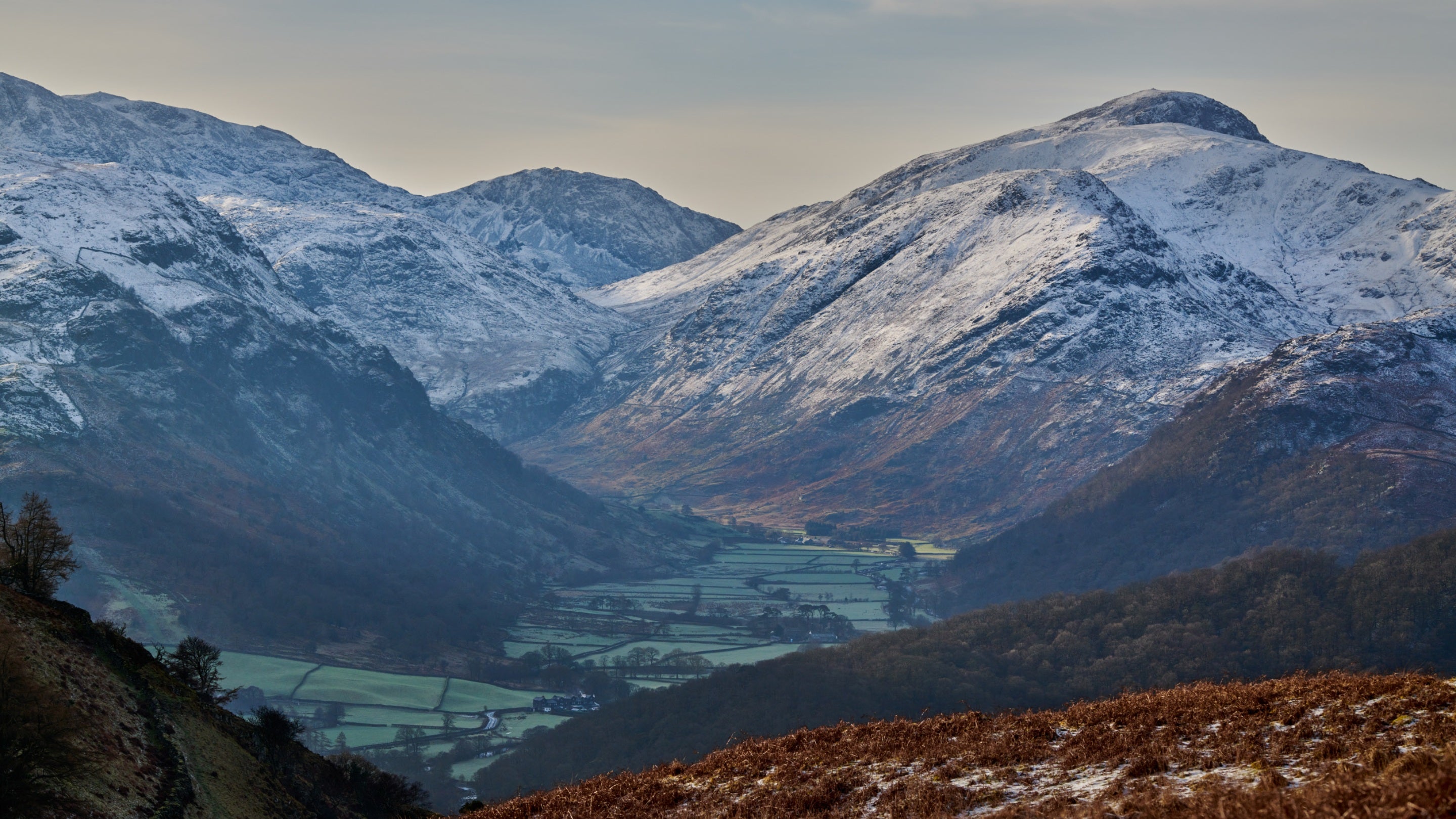 View of a green Lake District valley floor divided into small fields, with  snow-capped fells soaring around it