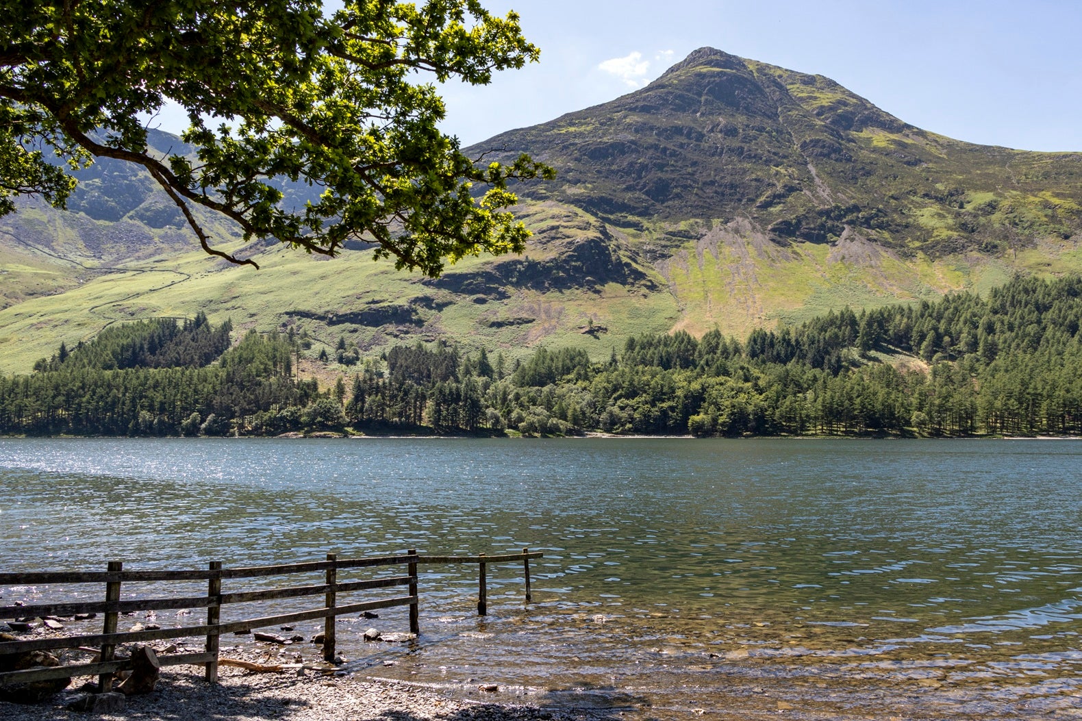 Lake with a partially submerged fence in the foreground, looking across to a sunlit green mountain and woodland beneath a clear blue sky.