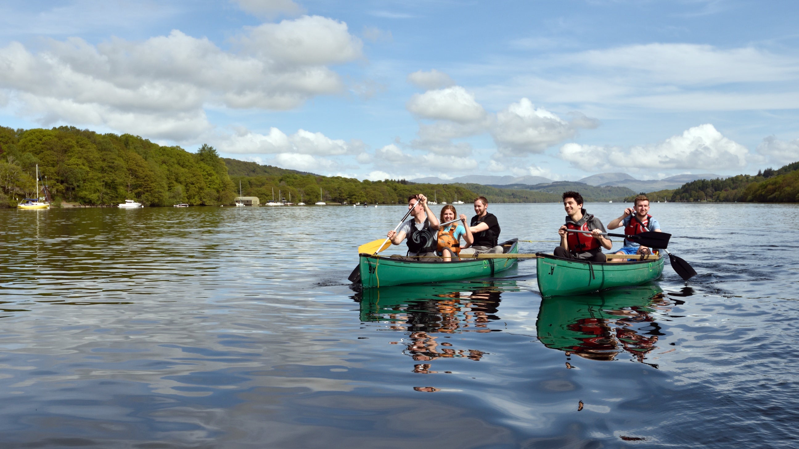 Two kayaks with people in on a reflective lake with vegetation and mountains behind  and blue skies with white clouds above.