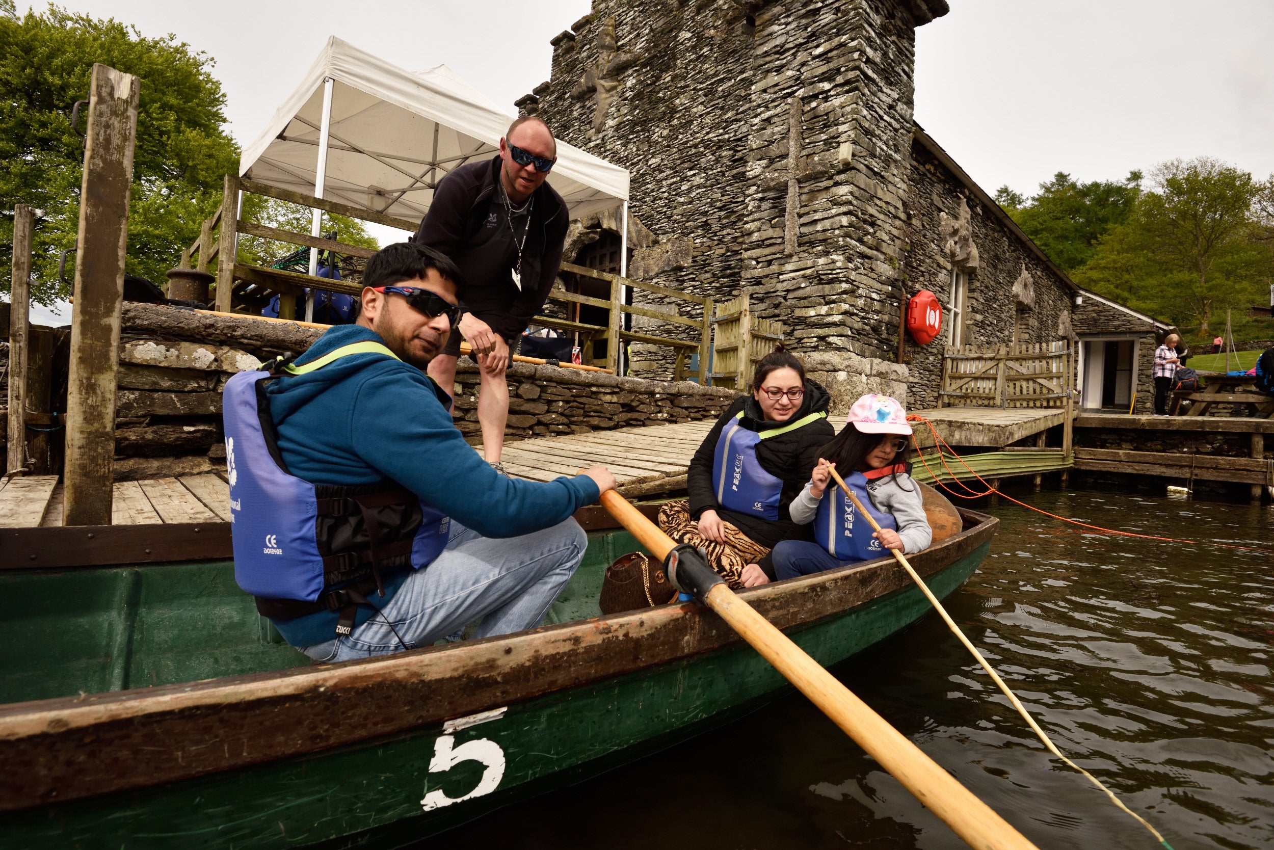 A family sat in a traditional wooden rowboat next to a stone jetty with a stone boathouse in the background.