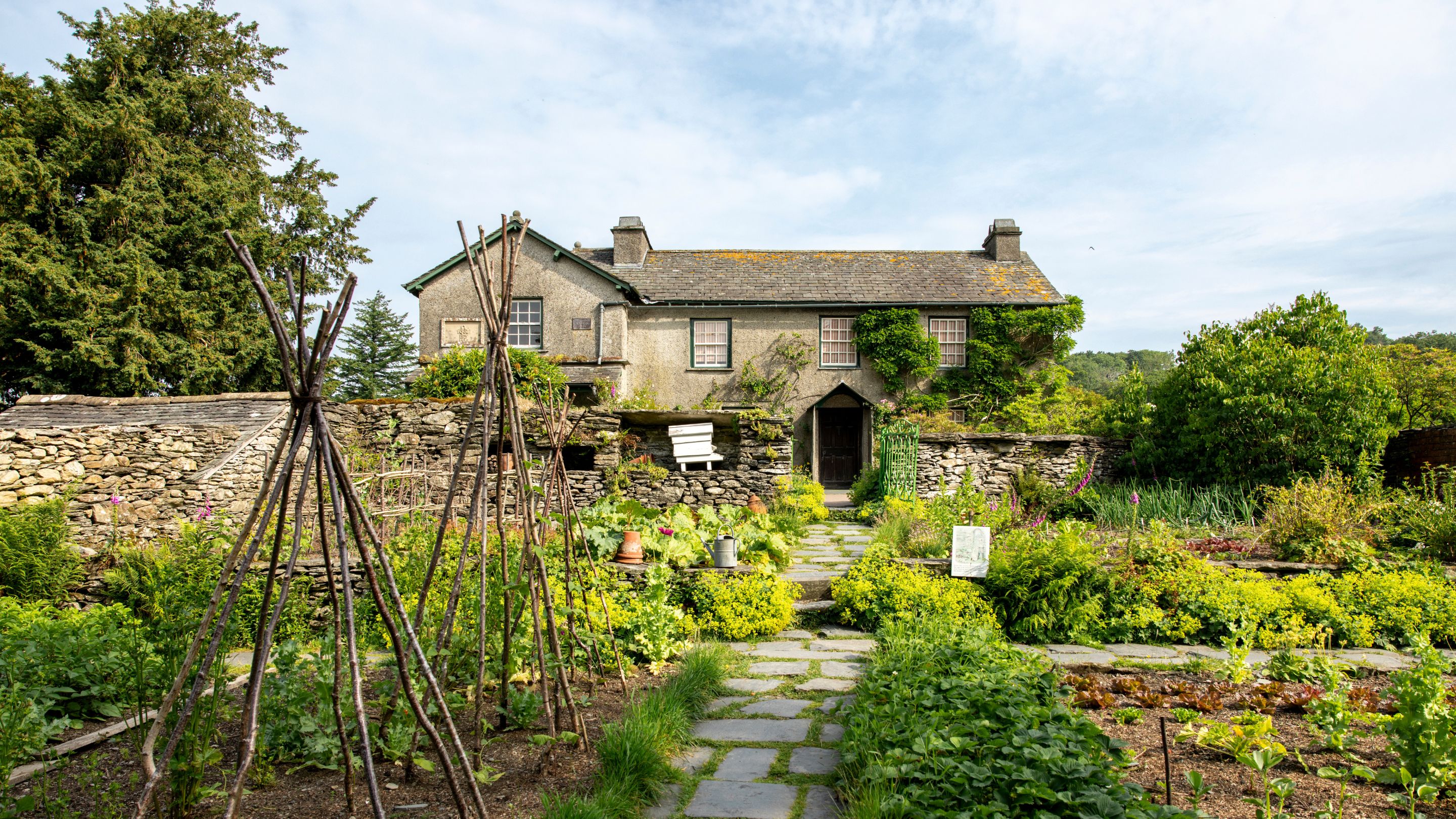 Vegetable garden at Hill Top in the Lake District, with stone paths, wooden plant supports and leafy beds leading to a traditional farmhouse behind a stone wall