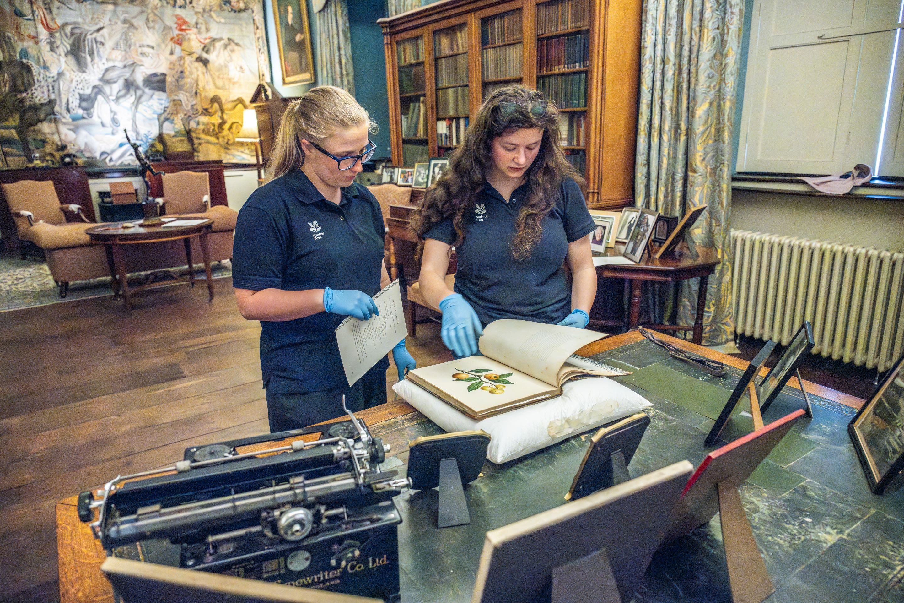 Two National Trust staff members, wearing navy blue polo shirts with the NT oak leaf logo, carrying out conservation work on a large, old book.