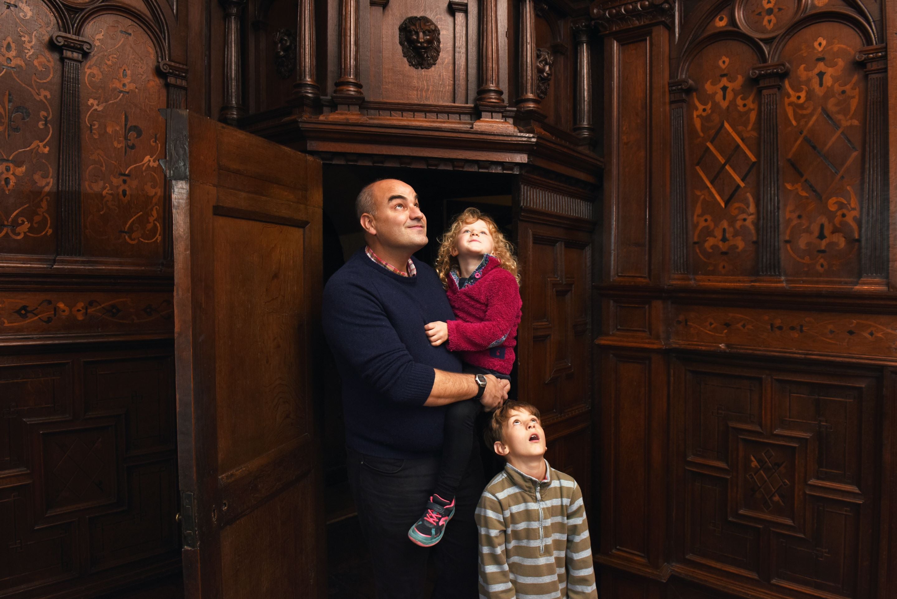 Father holding a toddler, with a small boy standing next to him. They are looking upwards at the elaborate wooden carvings in the house at Sizergh.