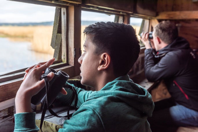 Young bird watcher and NT ranger sitting in the bird hide at Park End Moss wetland. They each have a pair of binoculars.