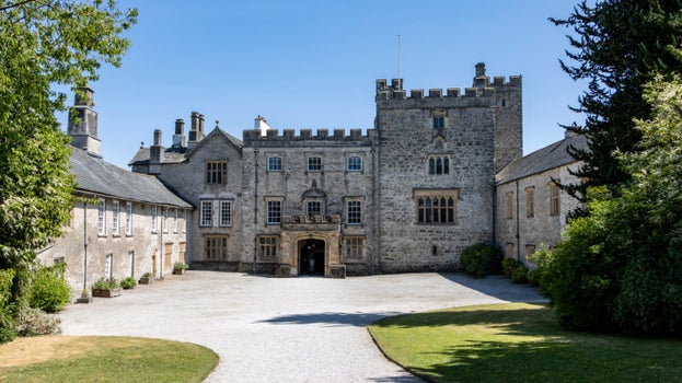 A courtyard with stone buildings on three sides. One side has crenellations and a solid square tower