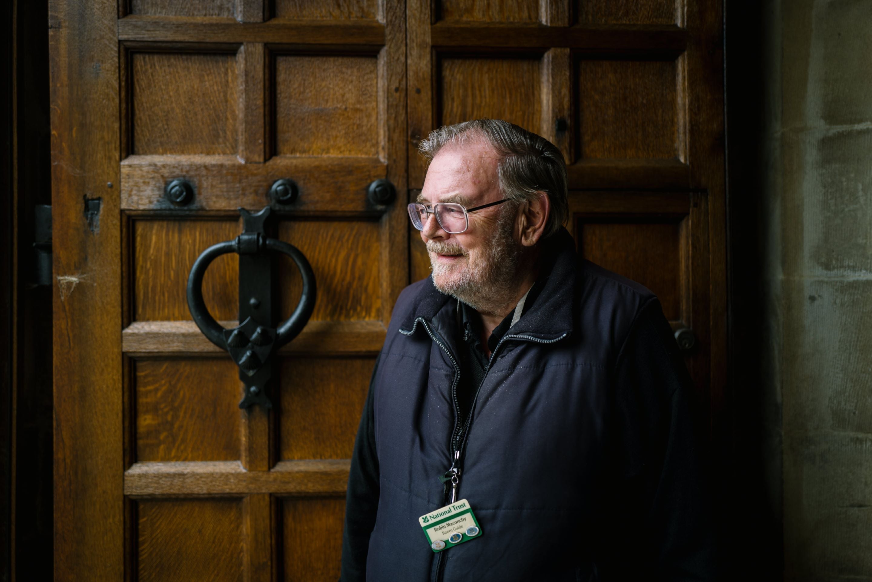 Male volunteer in glasses and a gilet, wearing a black volunteer lanyard and a name badge. He is waiting to greet people coming into the manor house at Sizergh, and stands in front of the large, dark oak door with it's black door knocker.