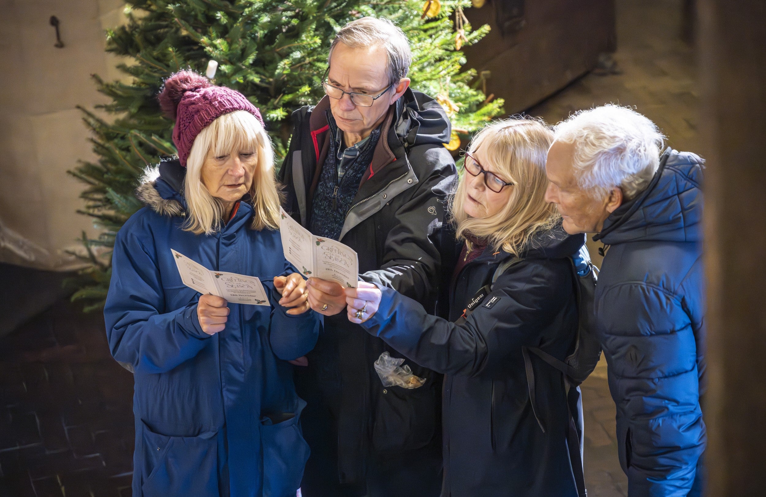 Four visitors, two older men and two older women, in winter coats and hats, in the entrance hall at Sizergh. They are reading their Christmas invitations.
