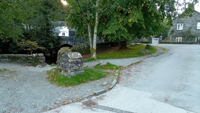 Car park entrance with a drystone wall and a road with a stone bridge in the background, Elterwater, Cumbria