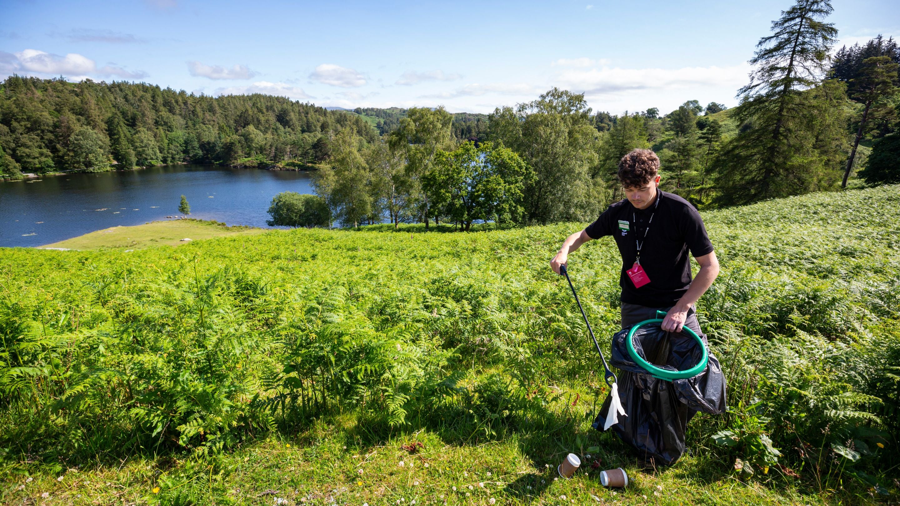 Volunteer litter picking at Tarn Hows, Cumbria. Behind him is a view of the lake and surrounding greenery.
