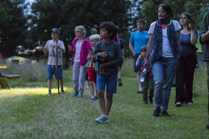 Group of families with children of different ages holding bat detectors in a field at dusk.