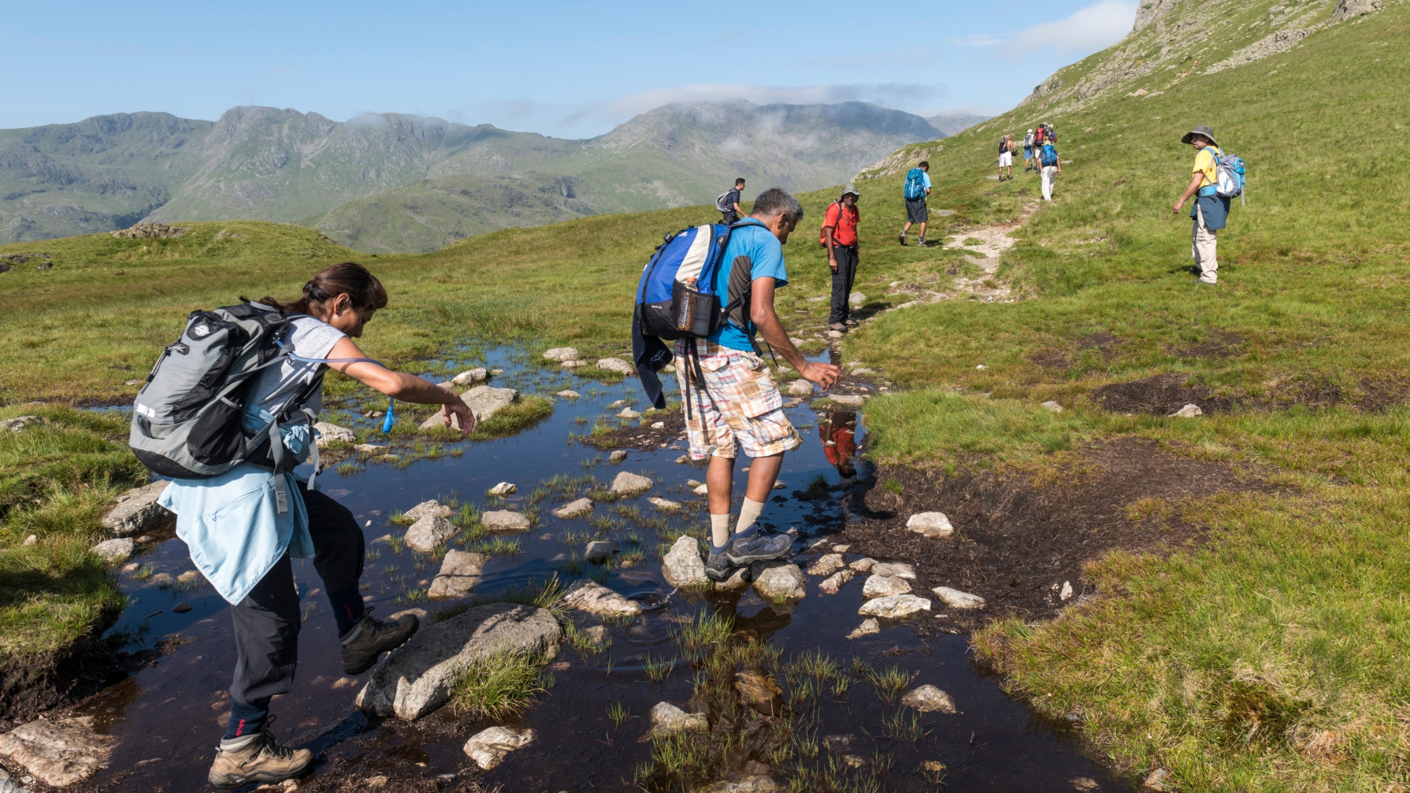 A group of walkers crossing stepping stones through a bog on a sunny Lake District fell