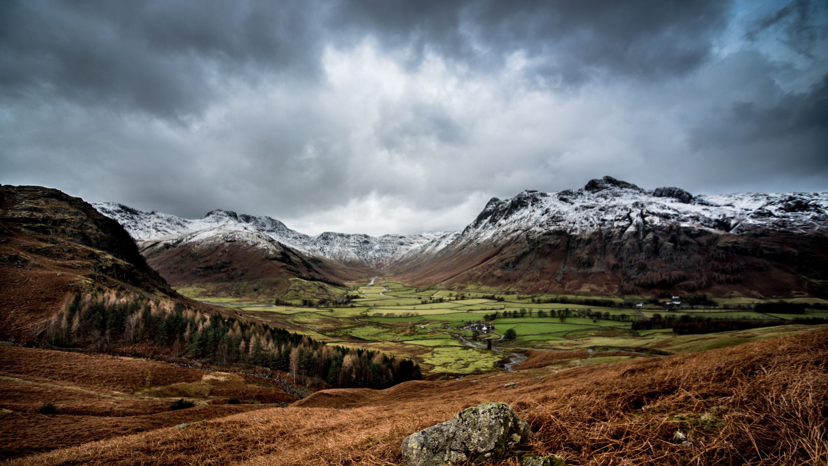 A dramatic landscape with orange bracken and green fields in the valley, framed by snow capped mountains and dark grey sky.