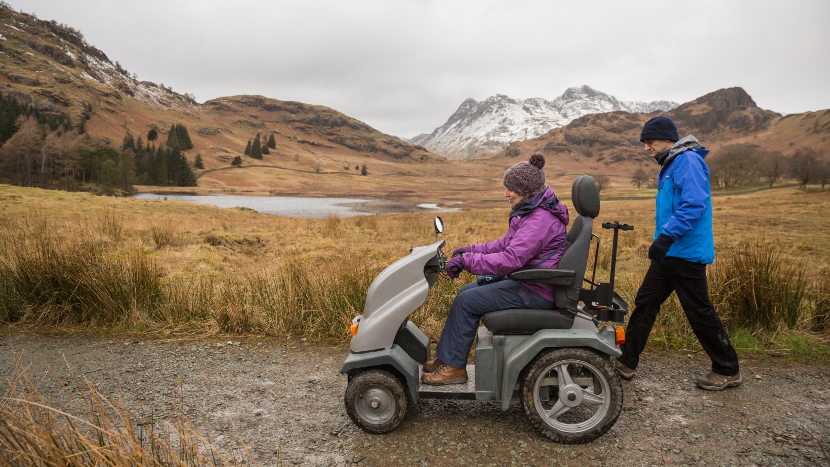 Two people in winter clothes exploring the snowy landscape around a tarn, one on foot and one on a tramper mobility vehicle