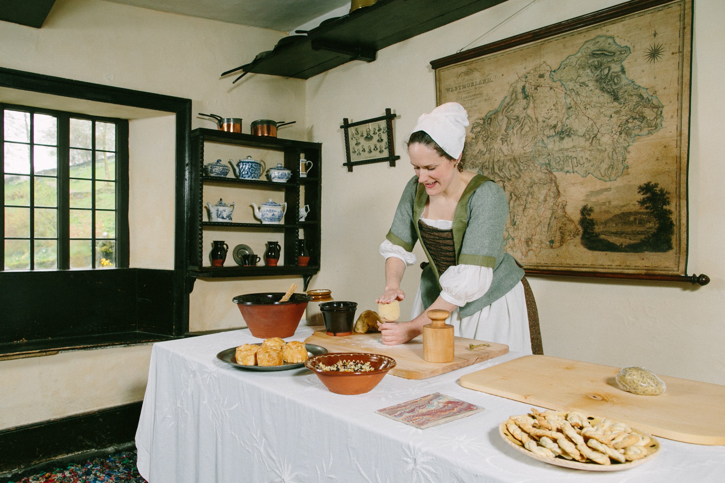 A woman in period costume creating food on a table in an old farmhouse kitchen