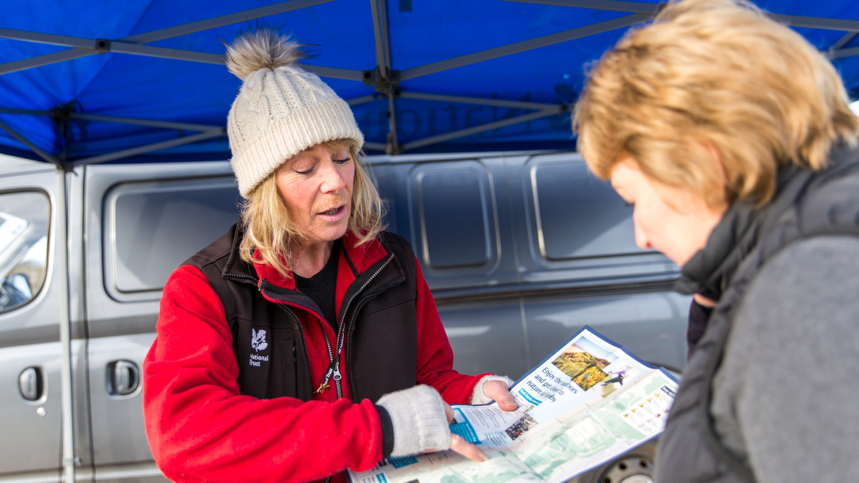 A member of the National Trust's Welcome Team, who is wearing winter clothing, talks to a visitor and presents them with a leaflet.