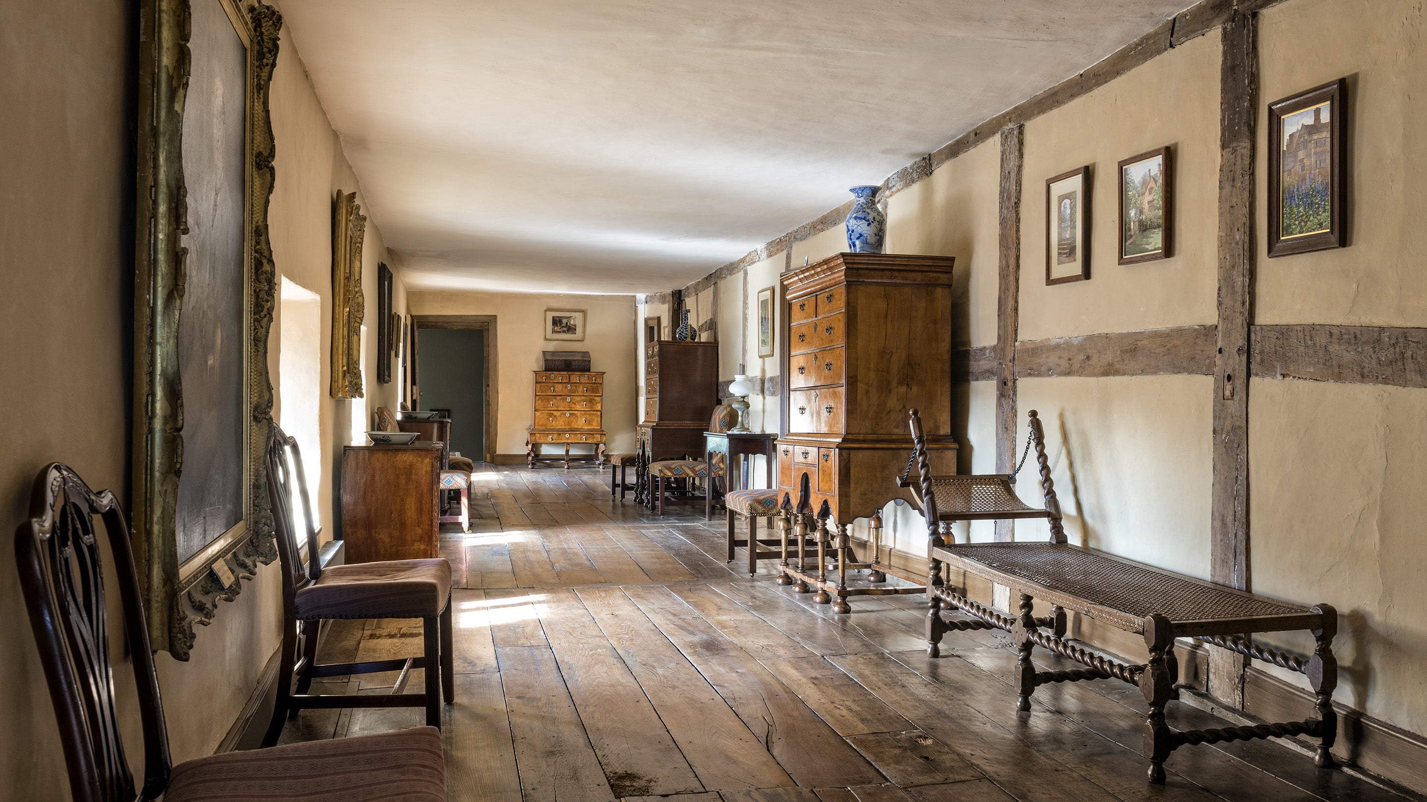A long, low ceilinged corridor with wide wooden floorboards and half-timbered walls. Antique furniture and paintings line the walls.