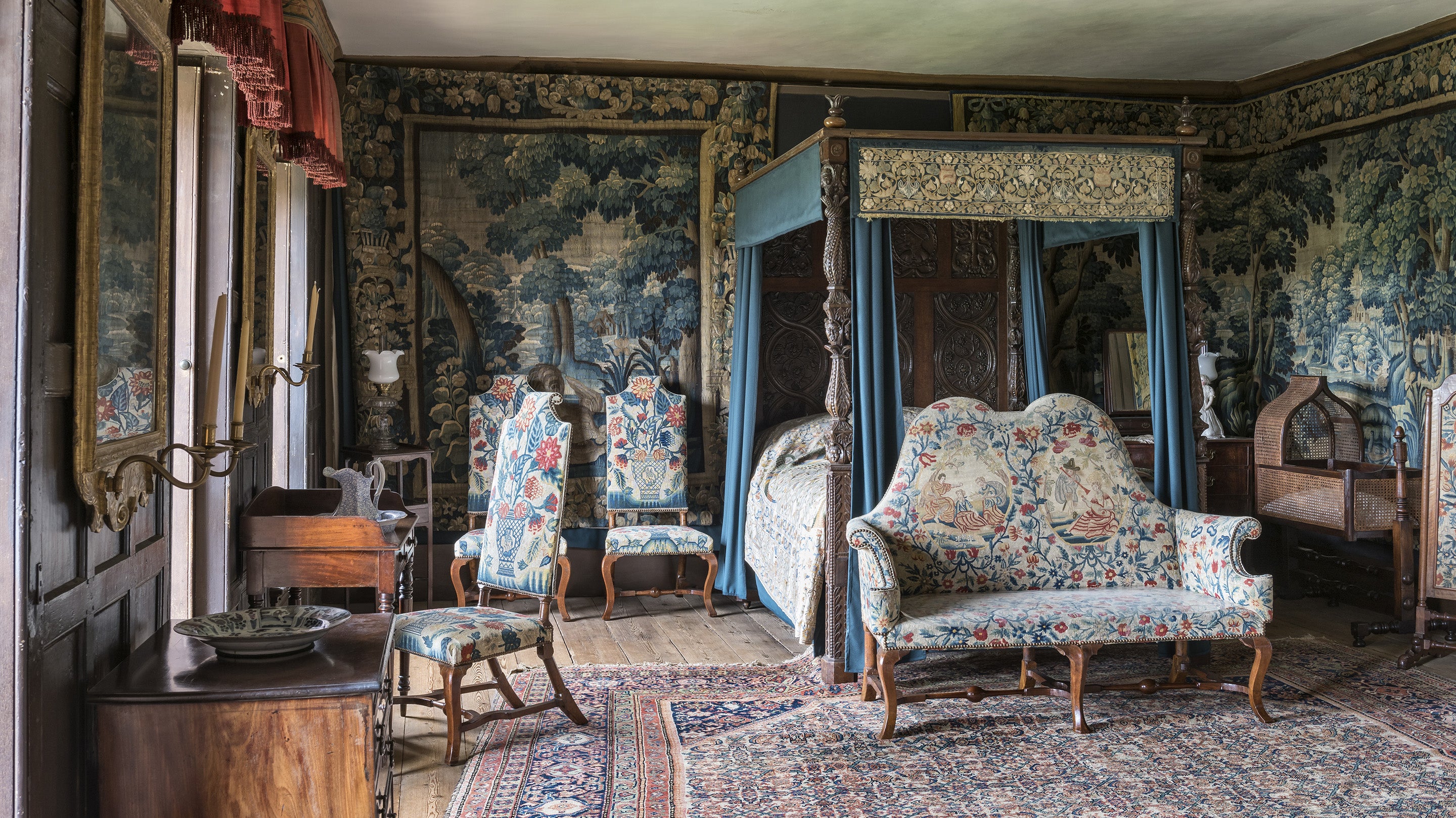 An elaborately decorated bedroom, with a four-poster bed, crib, three chairs, and a settee. The chairs and settee are upholstered in a decorative floral tapestry and there are tapestry wall hangings depicting trees.