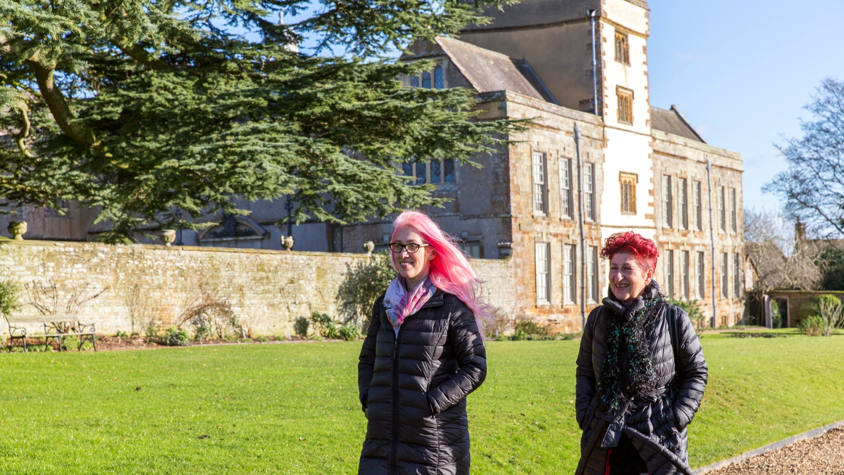 Two visitors in winter wear walking along a path with part of Canons Ashby house in the background